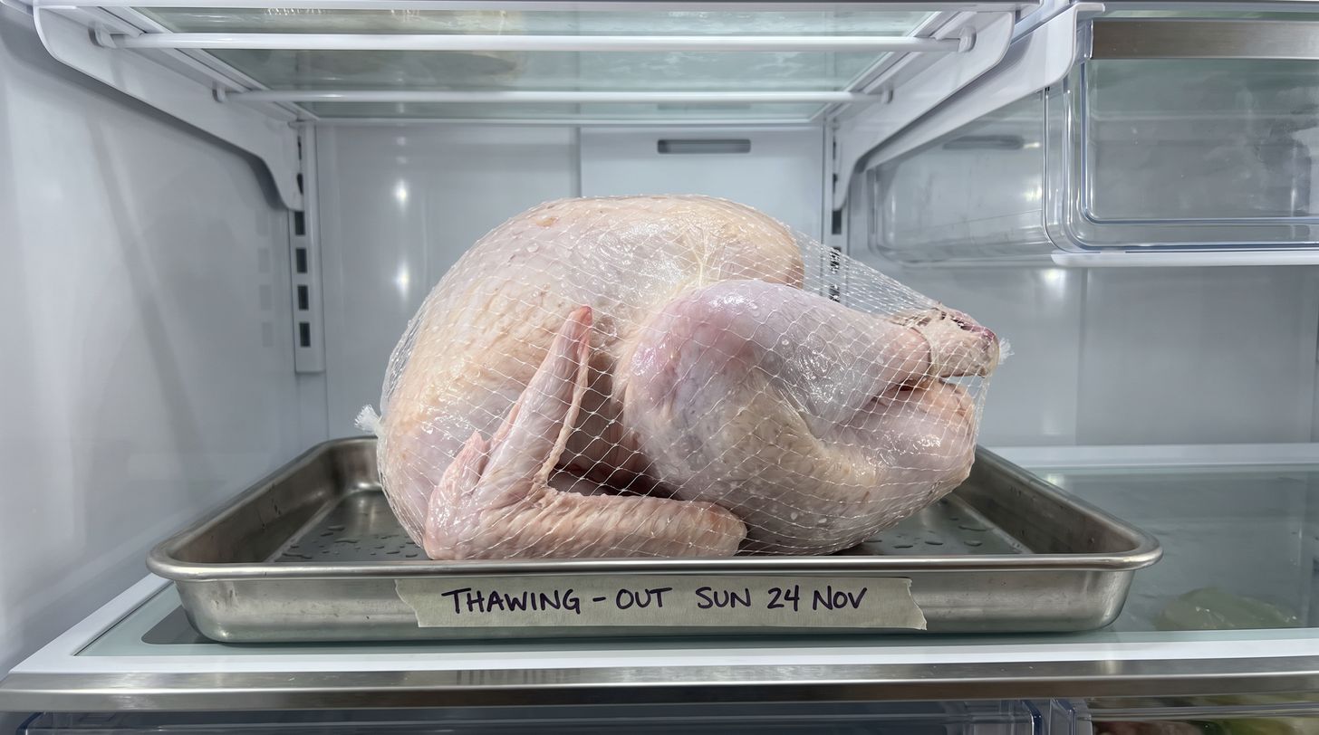 A whole raw turkey in netting thawing on a stainless tray in a refrigerator, with tape on the tray edge reading THAWING - OUT SUN 24 NOV